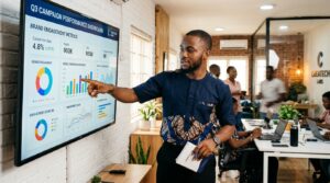 A young man in a creative agency office, standing in front of a large wall-mounted screen displaying brand engagement metrics and a campaign performance dashboard.
